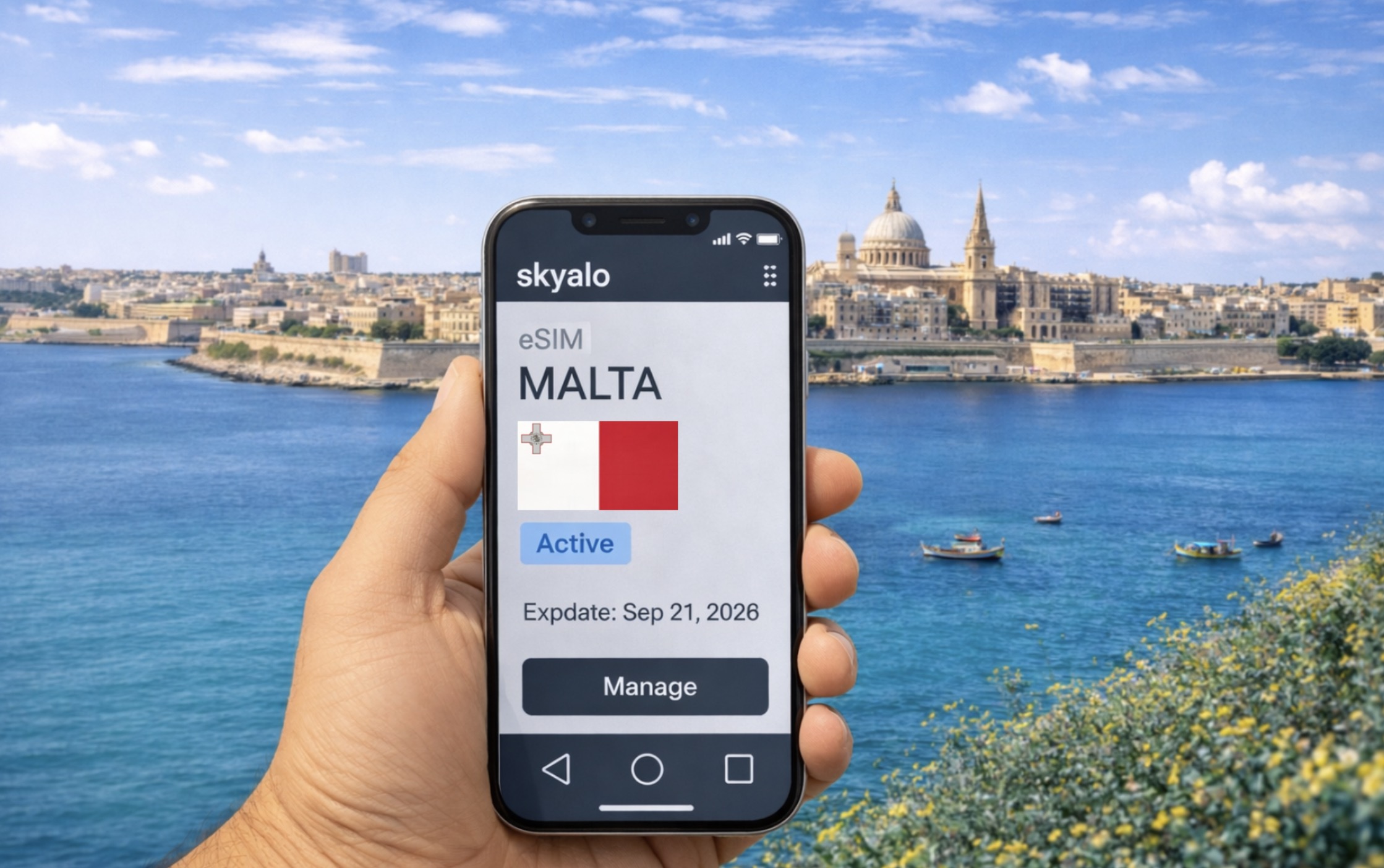 Telefon pintar dengan aplikasi Skyalo dan eSIM Malta yang telah diaktifkan di latar pantai Valletta yang cerah, laut biru dan bot tradisional.